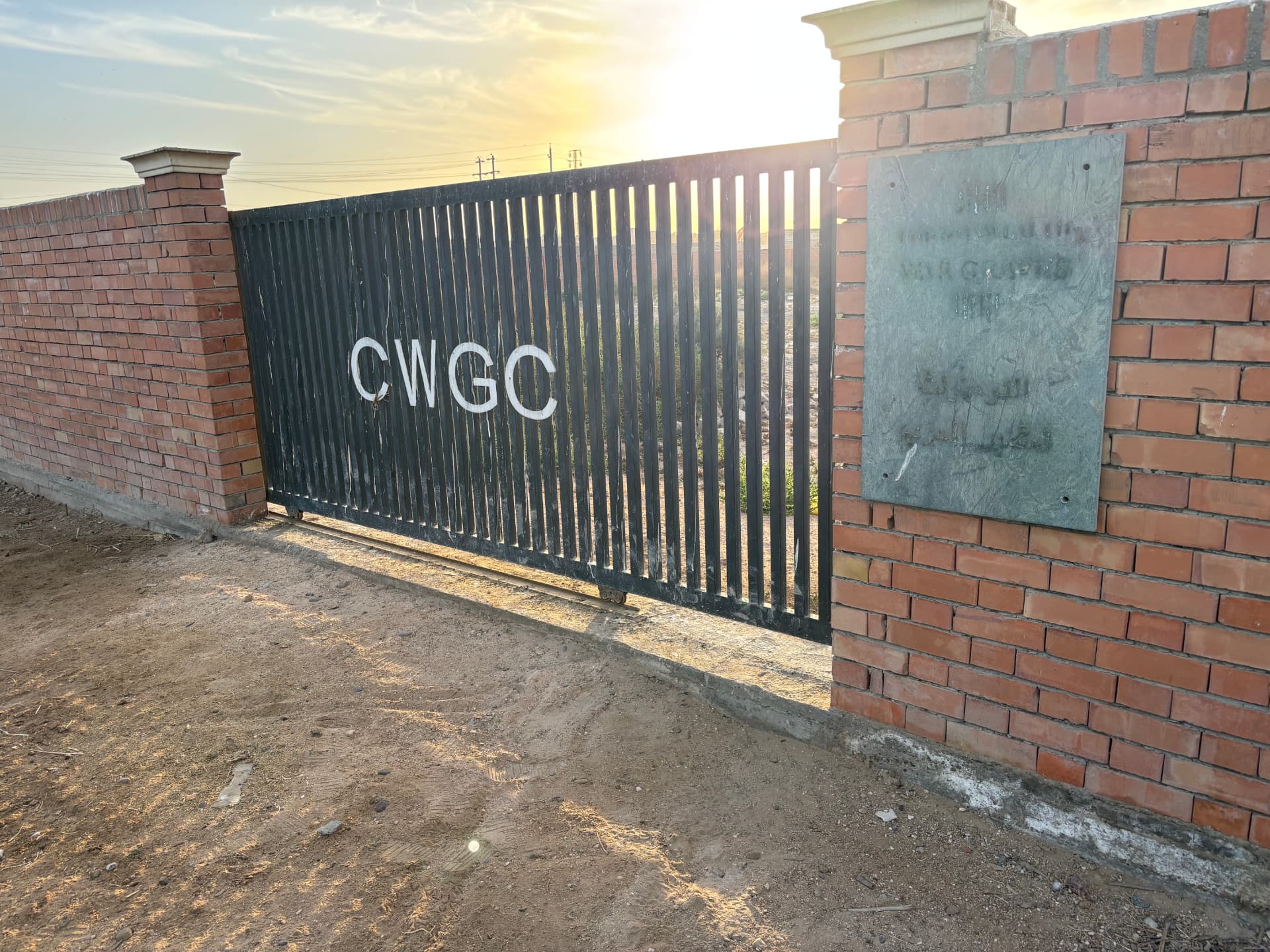 The entrance to Ma'asker al Raschid, the CWGC gate and boundary wall with the Commission's bilingual plaque beside it.