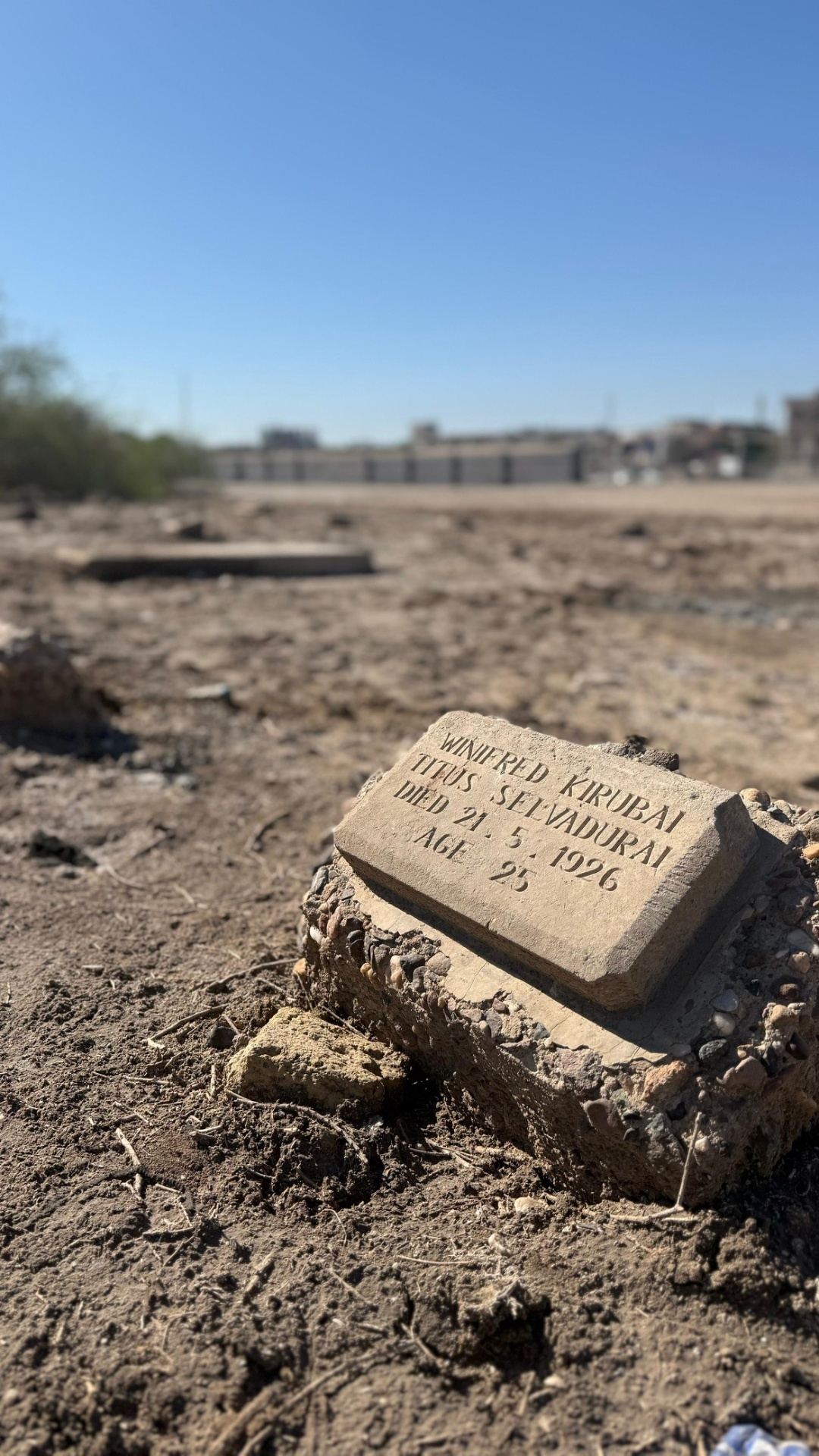 A small concrete headstone for Winifred Kirubai Titus Selvadurai (died 21.5.1926, age 25) standing alone on bare, sun-baked ground in the civilian section of Basra War Cemetery, with sparse vegetation and city buildings visible in the distance.