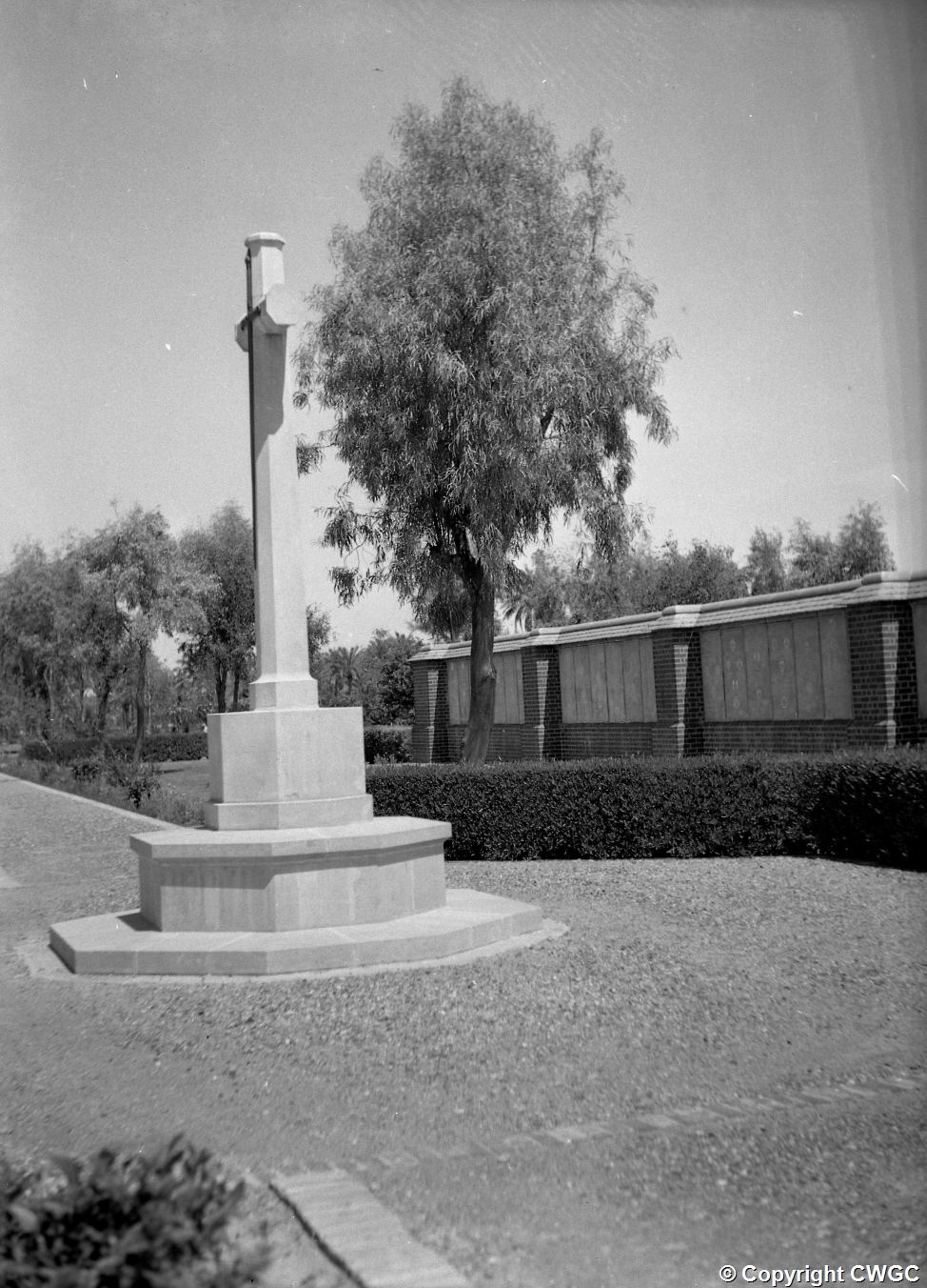 The Cross of Sacrifice standing on its tiered pedestal at the head of the gravel path, with a section of the Memorial Wall and its inscribed panels visible to the right beneath the trees. © CWGC.