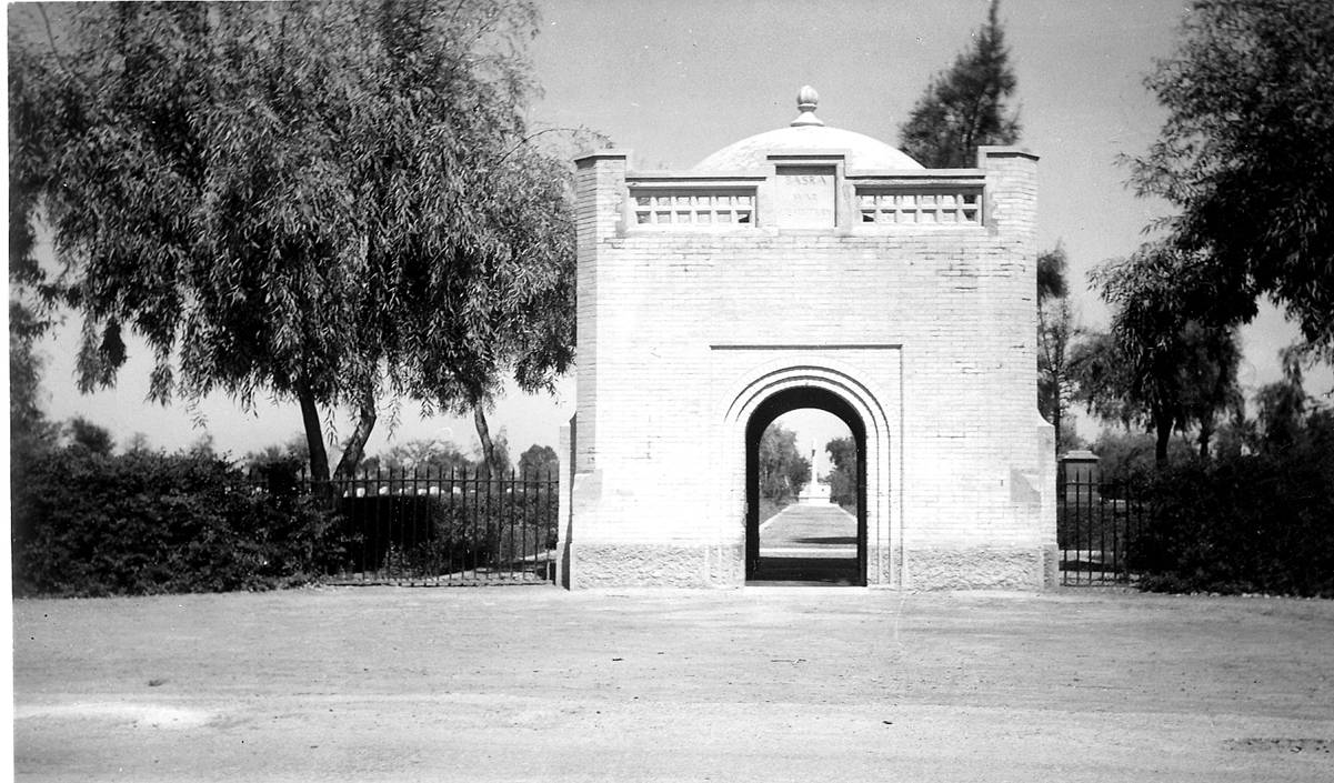 The domed gatehouse at the entrance to Basra War Cemetery, its arched passage framing a view down the central avenue towards the Cross of Sacrifice in the distance, with mature trees and iron railings to either side. © CWGC.