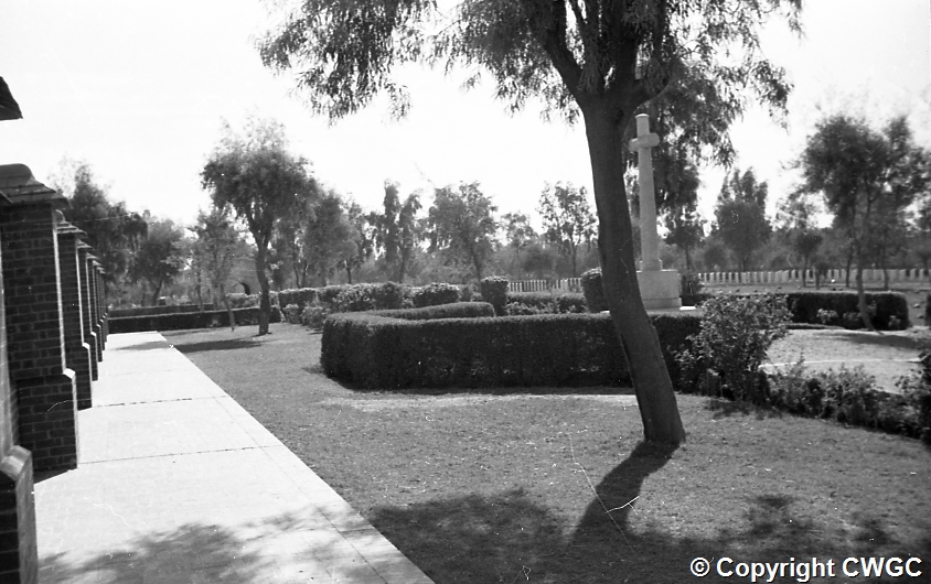 Looking along the Memorial Wall towards the Cross of Sacrifice in the middle distance, with clipped hedges, gravel paths and headstones visible at the far end of the cemetery. © CWGC.