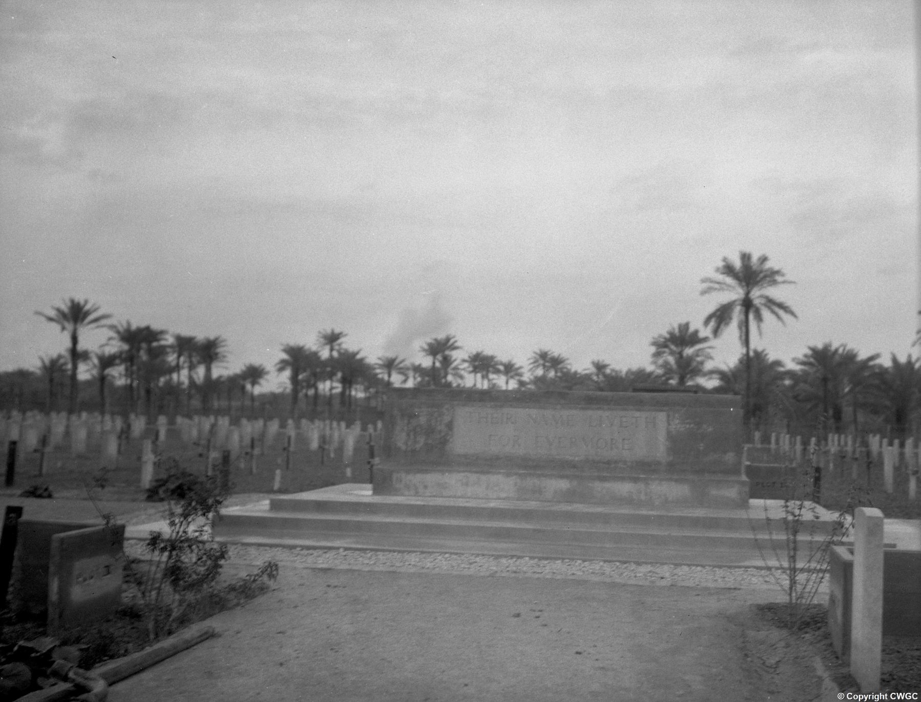 The Stone of Remembrance at Basra War Cemetery, circa 1920s/30s, standing on its tiered platform with rows of headstones and wooden crosses to either side and a backdrop of date palms.