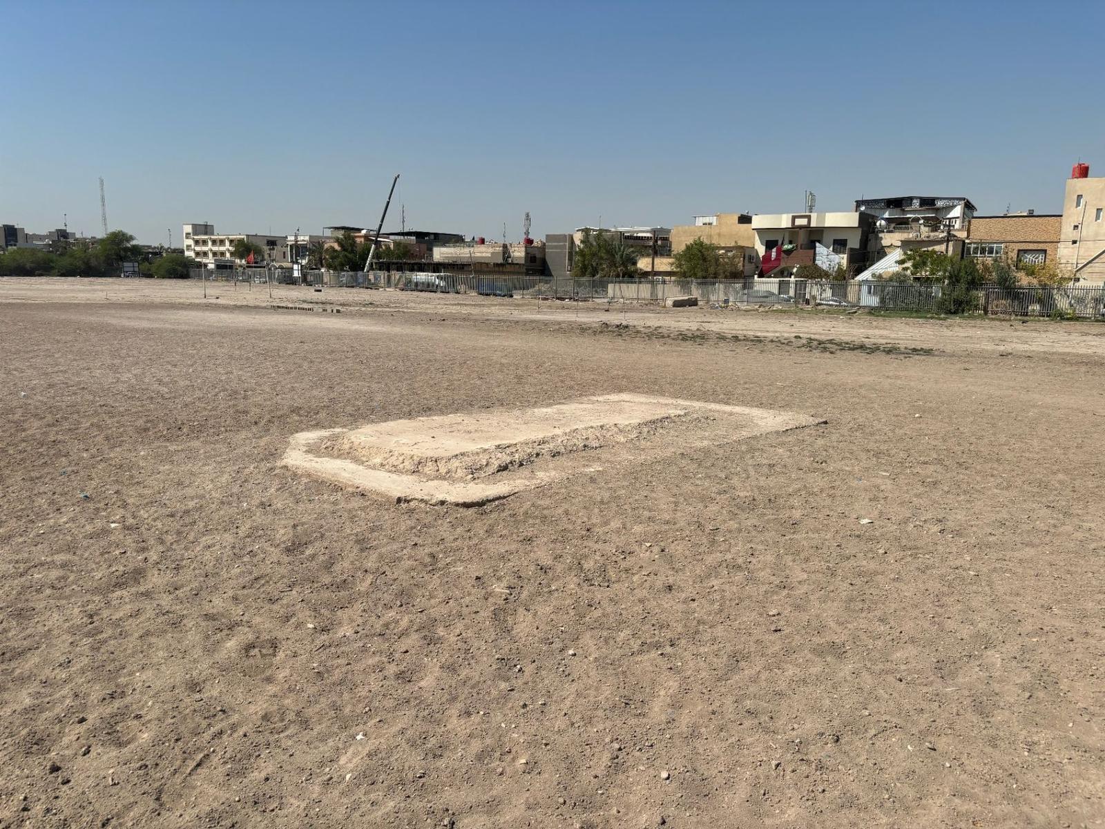 The empty plinth where the Stone of Remembrance once stood at Basra War Cemetery today, set in bare ground with modern Basra beyond the perimeter fence.