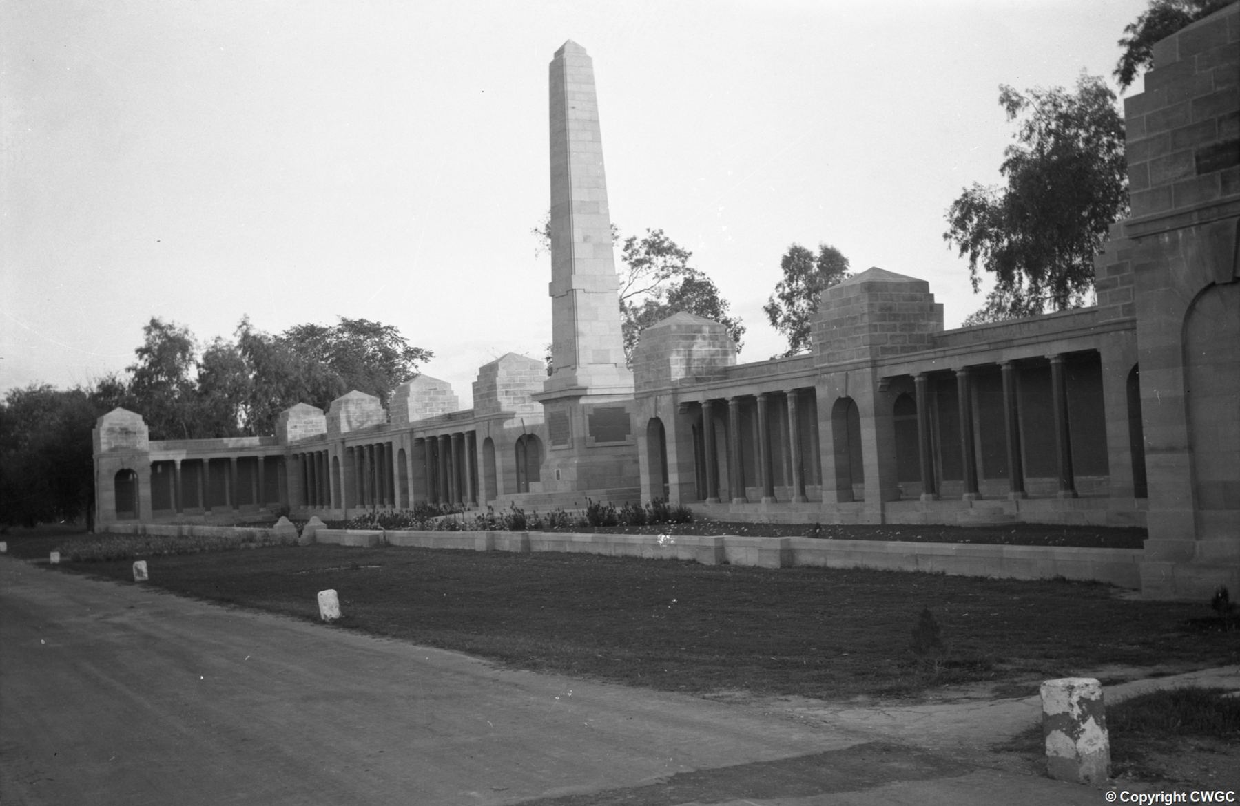 A wide view along the colonnaded screen wall of the Basra Memorial, with the central obelisk rising above the pavilions and the gardens laid out in the foreground. © CWGC.