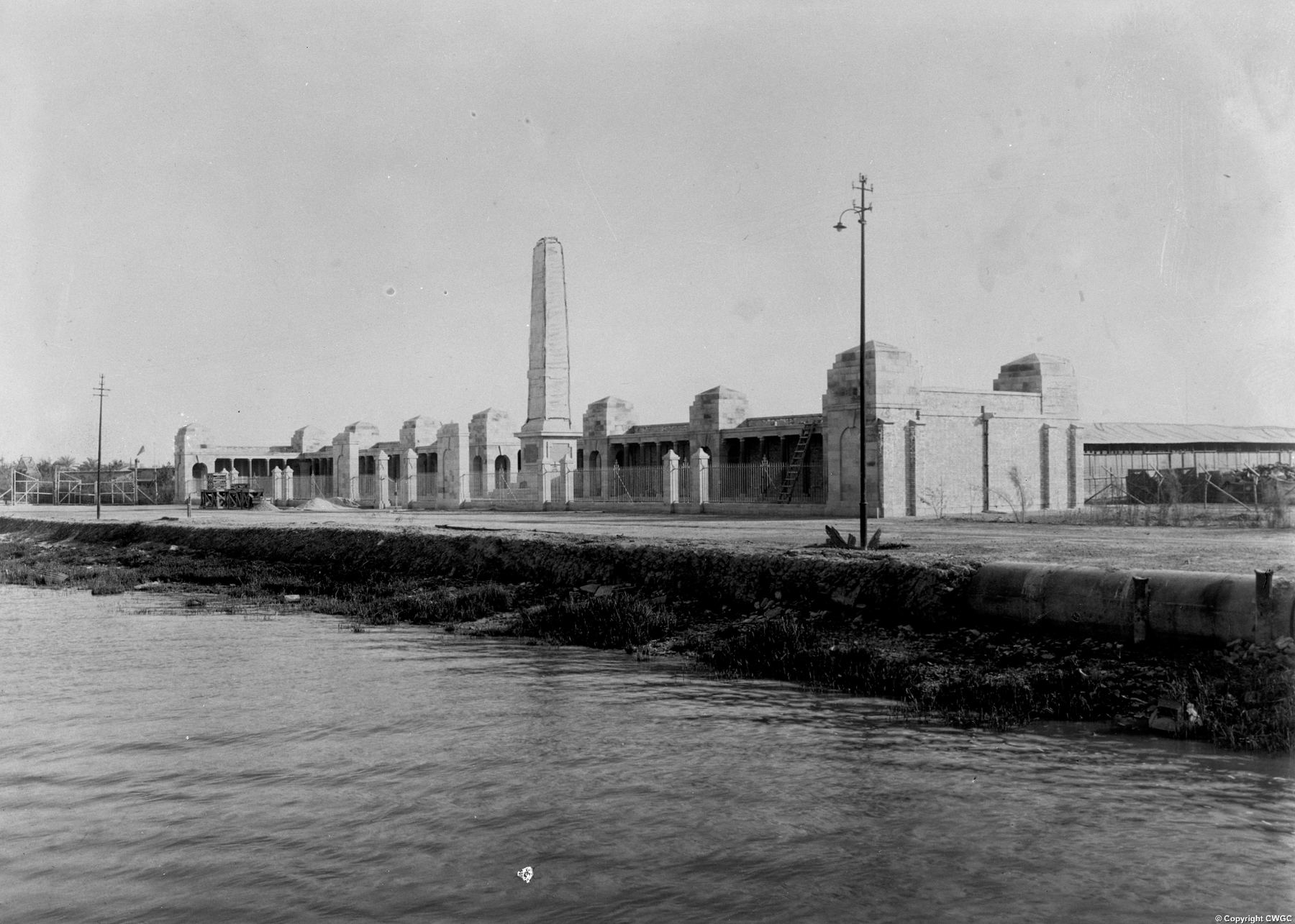 The Basra Memorial seen from across the waterway, with the obelisk and the full sweep of the colonnaded screen wall and pavilions visible along the bank. © CWGC.