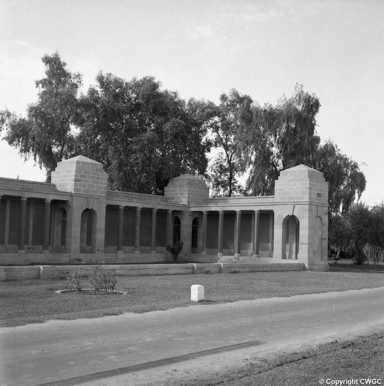 A side wing of the colonnaded screen wall, with its pavilions, arched openings and stepped entrance set behind a strip of lawn and garden borders. © CWGC.