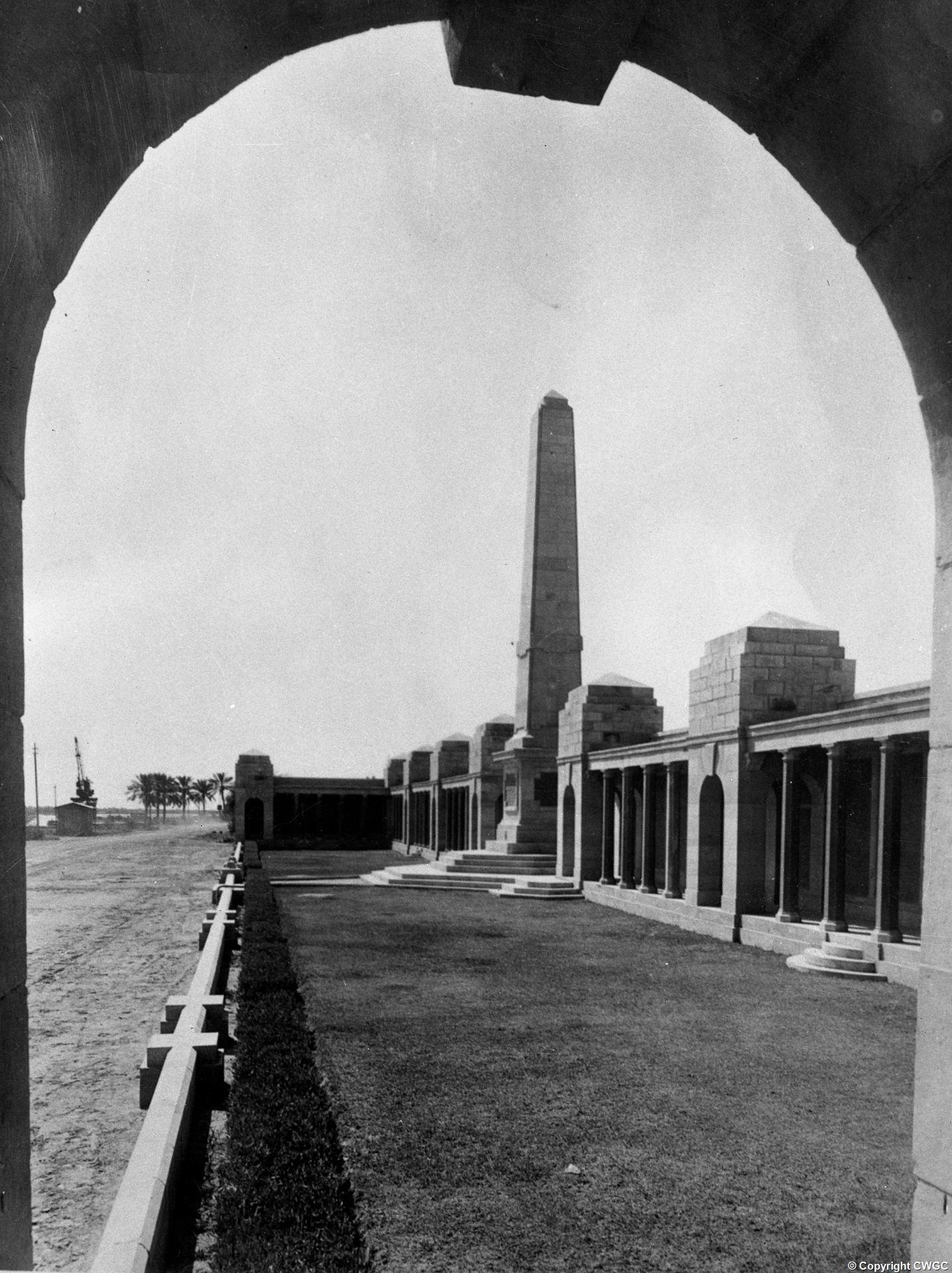 The Basra Memorial framed through one of the arched openings of the colonnade, looking towards the central obelisk with the Shatt al-Arab waterfront and a dockside crane visible at far left. © CWGC.