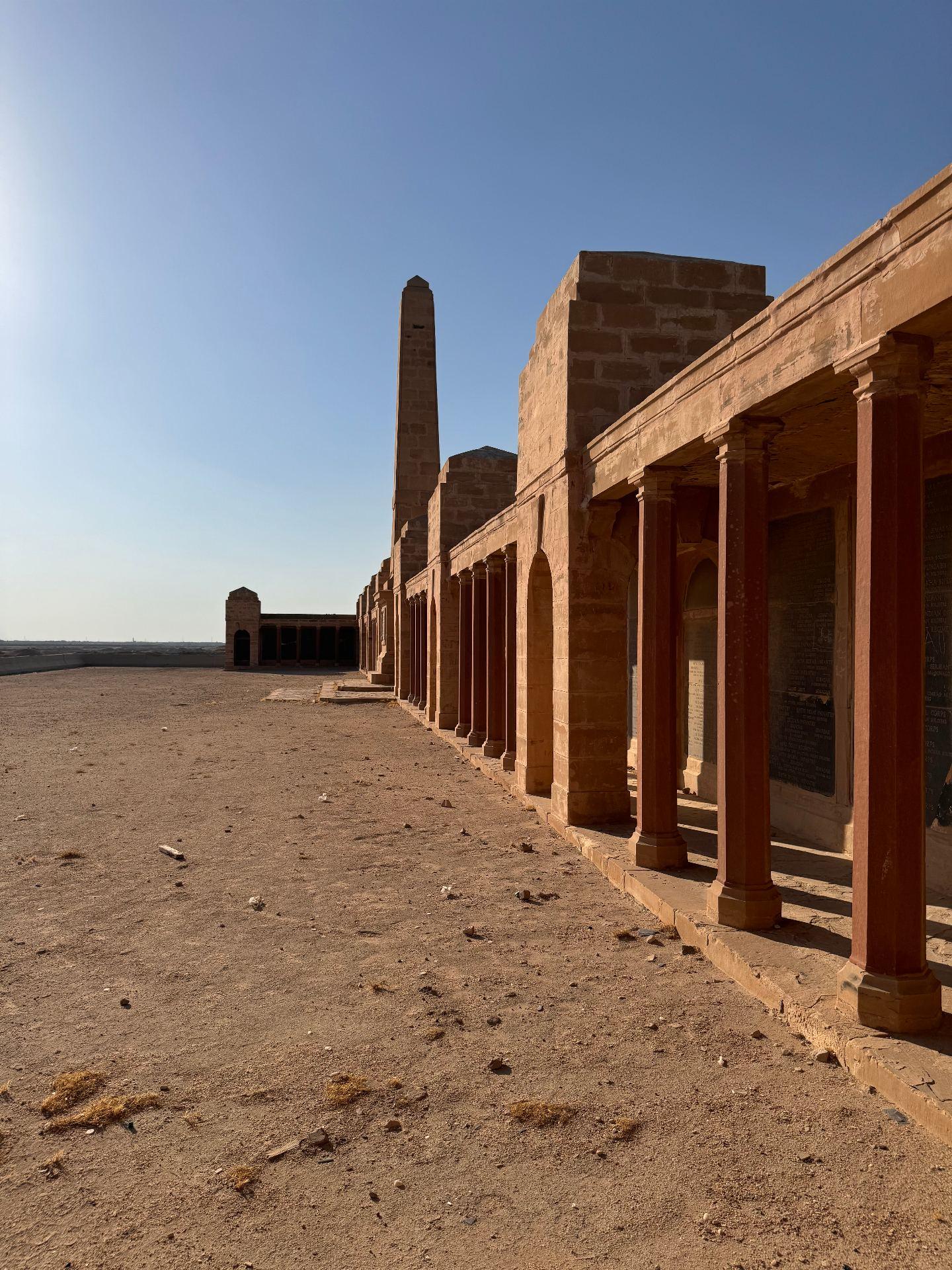 A close perspective view along the colonnaded screen wall of the Basra Memorial today, the inscribed panels visible behind the red sandstone columns, with the obelisk rising at the centre and a pavilion in the distance, all standing on bare desert ground.