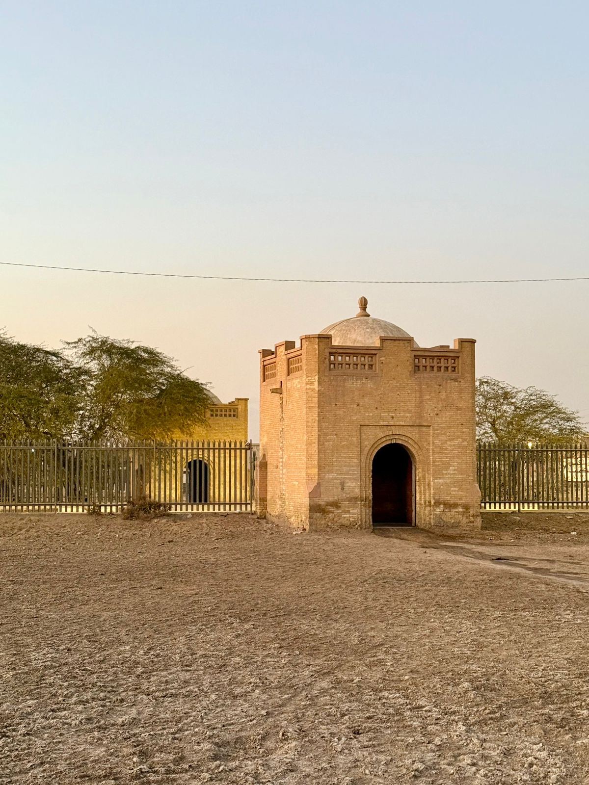 The two domed brick gatehouses of Basra War Cemetery seen from within the grounds at sunset, set behind the modern perimeter railings on bare, salt-scarred ground.