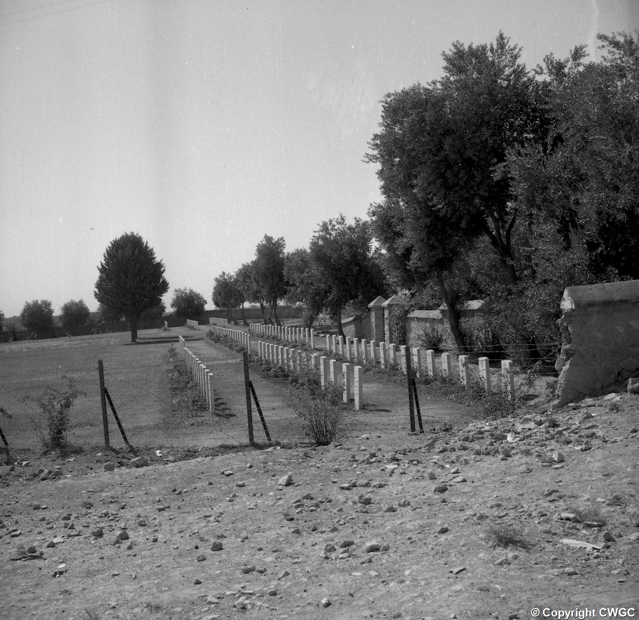 A view along the boundary showing long rows of headstones and damage to the perimeter wall. © CWGC.